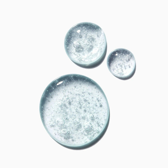 Close-up of Kérastase Genesis Homme Bain De Masse Épaississant Shampoo texture with clear, bubbly liquid drops on a white background